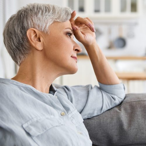 Side view of stylish elderly mature woman with neat make up and short hair relaxing on sofa at home, having thoughtful dreamy facial expression. Retired female sitting on couch in living room